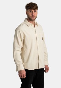 Camisa de pana beige claro con cuello abotonado, mangas largas y un bolsillo en el pecho. Presenta textura acanalada y una marca sutil.