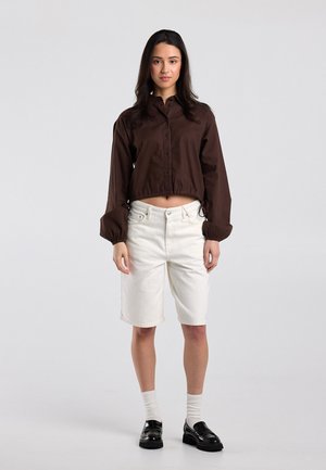 Jonge vrouw staand, draagt een donkerbruine cropped longsleeve, witte knielange shorts, witte sokken en zwarte loafers, op een lichtgrijze achtergrond.