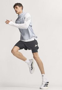 Athlet trägt leichte Weste, weißes Langarmshirt, schwarze Shorts und weiße Laufschuhe im Mittel-Schritt vor einfarbigem Hintergrund.