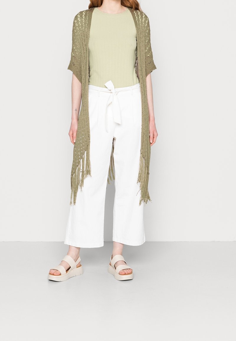 Cardigan ouvert en tricot vert olive avec des franges, associé à un haut côtelé vert clair et à un pantalon large blanc. Les sandales sont beiges avec une plateforme.