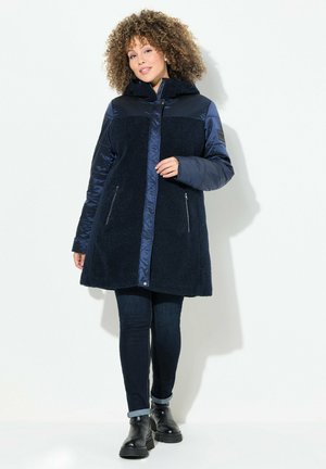 Navy blue coat with a plush textured body, shiny nylon sleeves, a hood, zippered pockets, and a buttoned front closure.