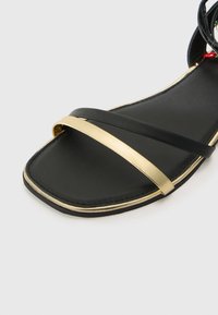 Sandalia plana negra con dos tiras delgadas negras y un acento de metal dorado. La suela es de color negro texturizado con un borde dorado delgado.