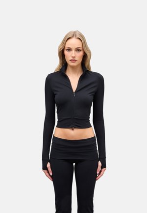 Donna con capelli biondi lunghi indossa un top corto nero aderente con cerniera e pantaloni neri a vita alta, in piedi contro uno sfondo chiaro e semplice.