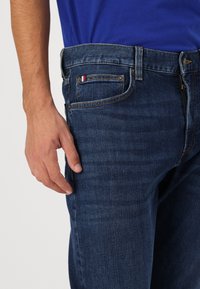 Dunkelblaue Denim-Jeans mit klassischer Passform, die eine subtile Textur und kontrastierende Nähte aufweisen. Enthält einen kleinen dreifarbigen Akzent auf der Tasche.