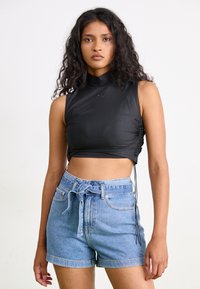 Zwarte, korte mouwloze top met een hoge kraag, gecombineerd met lichtblauwe denim shorts met een strik in de taille en opgerolde mouwen.