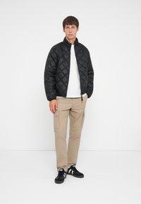Giacca nera trapuntata con colletto alto, indossata sopra una camicia bianca. Abbinata a pantaloni cargo beige e sneakers neri. Design semplice, senza motivi.