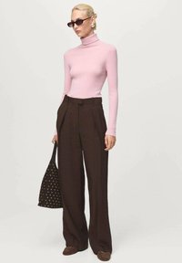 Femeie îmbrăcată cu un pulover cu guler înalt roz, pantaloni cu picioare largi de culoare maro închis, ochelari de soare negri, purtând o geantă mare negară cu detalii în formă de ochi.