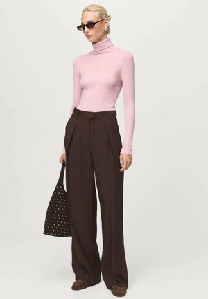 Femme portant un pull à col roulé rose, un pantalon large marron foncé, des lunettes de soleil noires, tenant un sac fourre-tout noir avec des détails en œillets.