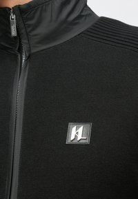 Juodas striukės modelis su užtrauktuku, turintis tekstūruotą megztinio kūną, lygų apykaklę ir sidabrinį logotipo pleistrą ant krūtinės. Pečiuose dekoruota ribbingais.