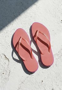 Paire de tongs rose Havaianas sur une surface blanche texturée avec un rayon de soleil et une ombre projetée en diagonale.