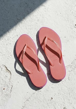 Egy pár rózsaszín Havaianas papucs texturált fehér felületen, amelyen a napfény és az árnyék átlósan vetül.