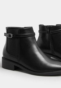 Svarta läderankelstövlar med en slät yta, utrustade med en dragkedja på sidan, spänndetalj och en låg blockklack.