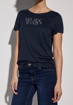 Frau trägt ein dunkelblaues T-Shirt mit dem Schriftzug "Nature & Revival" und dunkelblaue Jeans, steht vor einem schlichten hellen Hintergrund.