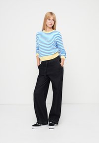 Modra in bela črtasta majica z rumenim robom, kombinirana s črnimi širokimi hlačami in črnimi supergami.