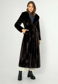 Dlouhý černý kabát z umělé kožešiny s širokým límcem, předními knoflíky a bočními kapsami, kombinovaný s robustními černými platformovými botami a krémovými kalhotami.