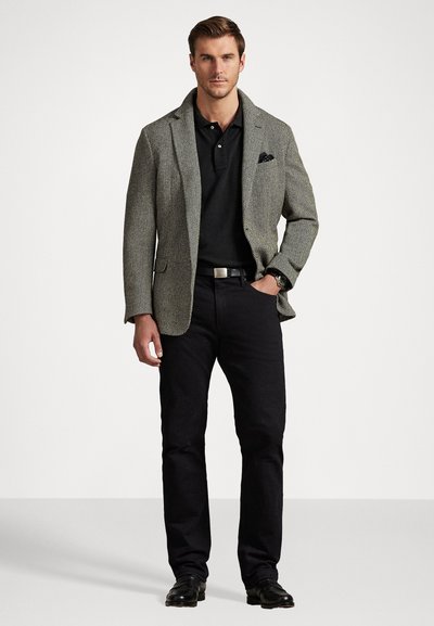Blazer cinza em padrão espinha de peixe sobre uma camisola pólo preta, combinado com jeans e sapatos pretos. Um lenço de bolso adiciona um detalhe ao blazer.