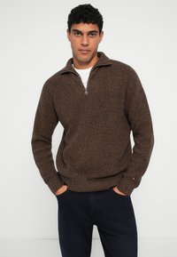 Brun lynlås sweater med et struktureret strikmønster, der har en stående krave og lange ærmer, parret med mørke bukser.