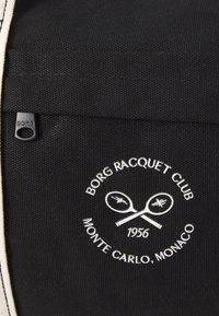 Fekete anyagú táska fehér varrással, kör alakú logóval, amelyen keresztbe tett teniszütők láthatók, valamint a "Borg Racquet Club, Monte Carlo, 1956" felirat.