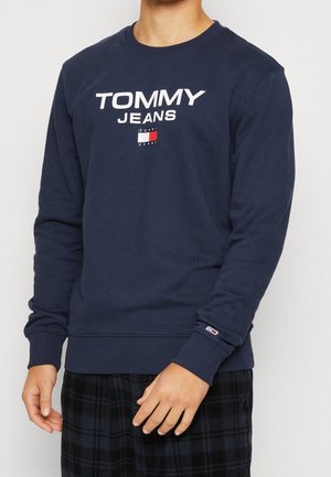 Mann trägt ein marineblaues Tommy Jeans Sweatshirt mit Logo und Flagge auf der Brust, steht mit entspannten Armen, von den Schultern bis zu den Hüften gezeigt.