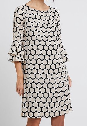 Vrouw draagt een zwart jurkje tot op de knie met grote witte polkadots en gerimpelde driekwart mouwen tegen een effen achtergrond.