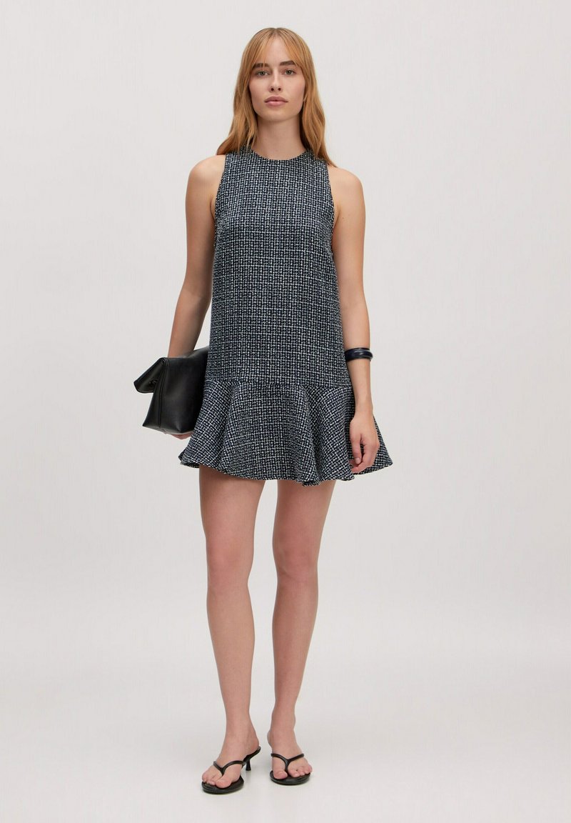 Ärmelloses schwarzes und weißes gemustertes Kleid mit einem ausgestellten Saum, aus strukturiertem Stoff. Kombiniert mit einer schwarzen Clutch-Tasche und minimalistischen Sandalen.