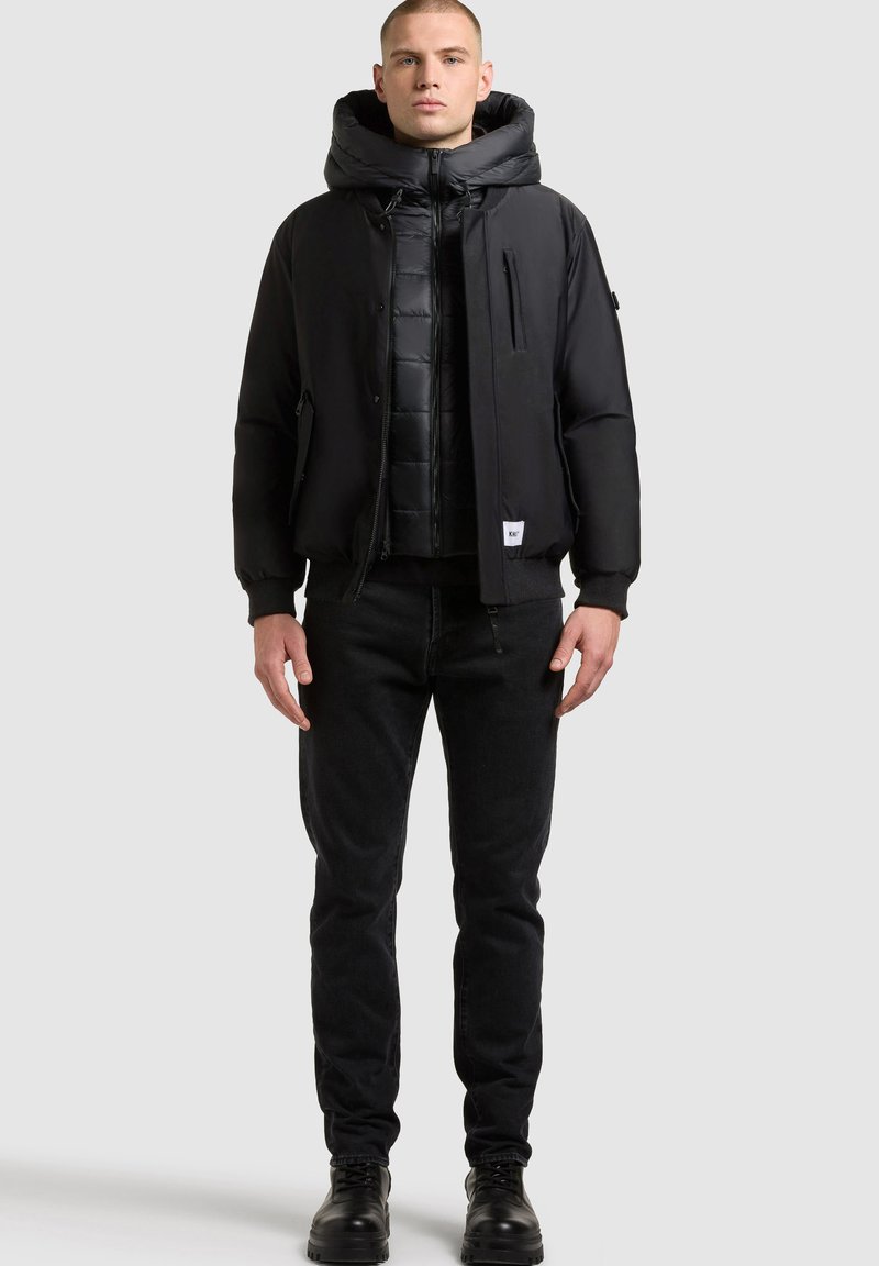 Chaqueta de plumas negra con capucha desmontable, cierre de cremallera y bolsillos laterales, superpuesta sobre una chaqueta interior negra acolchada y combinada con jeans negros y botas.