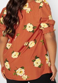 Kwitnący top w kolorze rdzawym, z żółtymi różami i zielonymi liśćmi. Krótkie bufiaste rękawy z marszczonymi elastycznymi mankietami. Miękka tkanina, luźny krój.