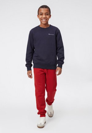 Lächelnder Junge, der läuft, trägt ein schwarzes Champion-Sweatshirt, rote Hose und weiße Turnschuhe vor einem schlichten weißen Hintergrund.