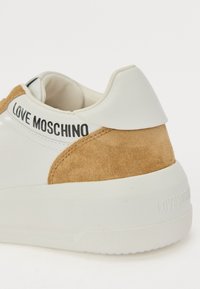 White leather sneaker with beige suede accents, rounded heel, and embossed "LOVE MOSCHINO" logo on the back, featuring a textured sole.