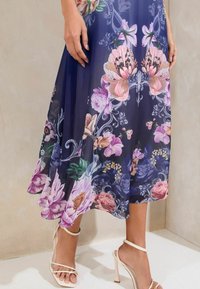 Vestido de gasa floral con base azul marino, que presenta grandes flores rosas y moradas, con un dobladillo fluido, y combinado con sandalias de tacón nude.
