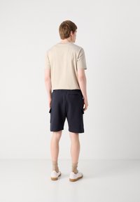 Beige pamut póló és sötétkék rövidnadrág oldalsó zsebekkel. A modell világos színű zoknit és fehér, gumi talpú cipőt visel. Egyszerű dizájn.