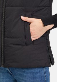 Schwarze gepolsterte Weste mit Reißverschluss, Seitentaschen und gerippten Strickbündchen. Der Stoff hat eine glatte Textur mit dezenten Steppdetails.