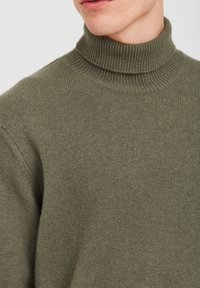 Maglione a collo alto verde oliva realizzato in morbido tessuto a maglia, con colletto e polsini a coste, caratterizzato da una trama liscia e un design aderente.