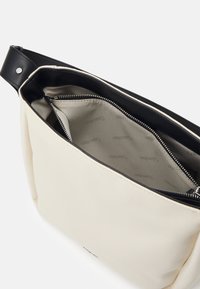 Sac à épaule en tissu couleur crème avec garniture en cuir noir, fermeture éclair ouverte sur le dessus et doublure intérieure beige imprimée avec le branding.