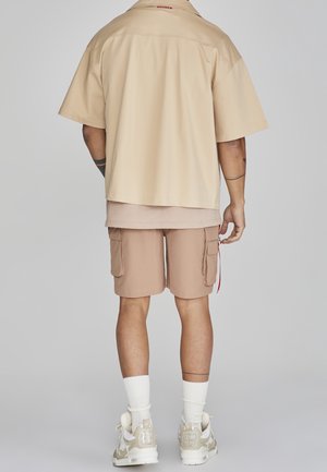 Chemise beige à manches courtes, superposée avec un haut beige clair, associée à un short cargo marron clair avec des poches, et des baskets de couleur claire.