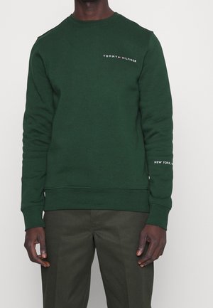 Mężczyzna w ciemnozielonej bluzie Tommy Hilfiger z logo na piersi i napisem „New York” na rękawie, w zestawieniu z ciemnozielonymi spodniami.