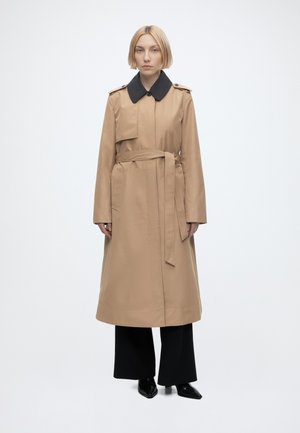Kvinde stående iført en beige trenchcoat med en sort krave, sorte vidde bukser og sorte spidse sko på hvid baggrund.