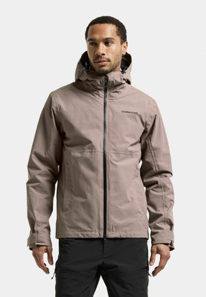 Man wearing a light brown hooded Didriksons jacket with black zipper and black pants, standing and looking slightly to the side.