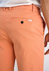 Pantalons en coton orange avec une poche arrière dotée d'un bouton noir, des poches latérales et une petite étiquette de marque sur la taille.