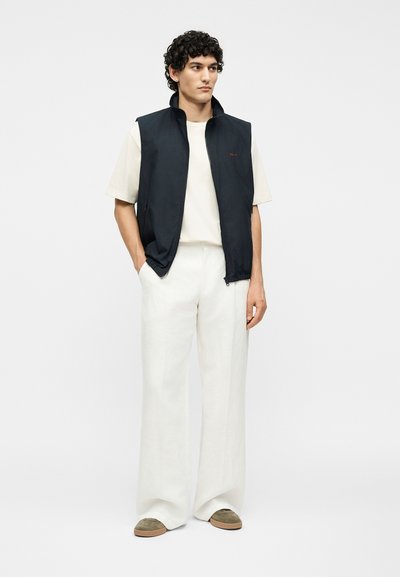 Νέος άνδρας με σγουρά μαλλιά, που φοράει μαύρο αμάνικο γιλέκο πάνω από λευκό μπλουζάκι και φαρδιά λευκή παντελόνια, με τα χέρια στις τσέπες.