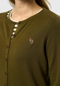 Olivgrön kofta i strukturstickad tyg, med rund halsringning, knappstängning och en liten broderad design på bröstet.