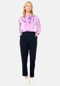 Lilac satīna blūze ar loku, apvienota ar sašaurinātiem melniem biksēm. Modelis valkā baltus sporta apavus, demonstrējot minimālistisku, elegantu apģērbu.