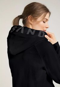 Schwarzer Kapuzenpullover aus strukturiertem Material, mit dem Wort "SAND" in fetten, kontrastierenden Buchstaben auf der Rückseite bedruckt.