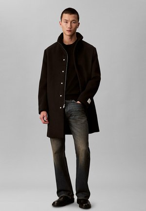 Junger Mann steht und trägt einen schwarzen Mantel über einem schwarzen Hemd, ausgewaschene schwarze Jeans und schwarze Schuhe vor einem einfarbigen grauen Hintergrund.