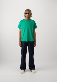 Grønn crewneck t-skjorte, løse mørke denimbukser og hvite sneakers. Modellen har på seg svarte solbriller og står foran en ensfarget bakgrunn.