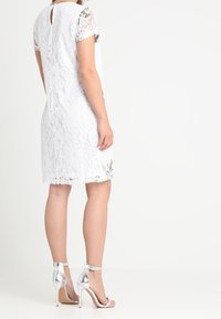 White lace dress with short sleeves, featuring a keyhole back and floral accents. Paired with silver high-heeled sandals.