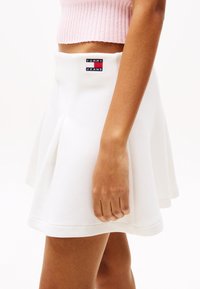 White skater skirt made from textured fabric, featuring pleats and a Tommy Jeans logo at the waistband. Close-up of hand on skirt hem.