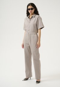 Ljusgrå jumpsuit med korta ärmar, figursydd midja, knappstängning och sidodetaljer; kombinerad med svarta pumps med spetsig tå.