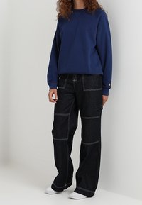 Osoba ubrana w niebieską bluzkę z długim rękawem, czarne szerokie spodnie z białymi przeszyciami i białe buty, stoi pod białą ścianą.