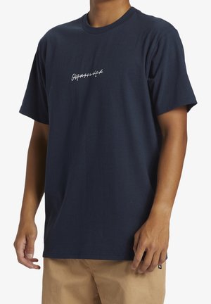 Osoba ubrana w granatowy T-shirt z małym białym przekreślonym tekstem na piersi i beżowe spodnie, stojąca na białym tle.